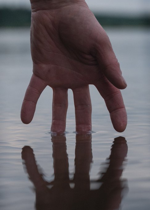 A close-up photograph of a hand reaching down to touch the surface of calm water, creating a reflection.