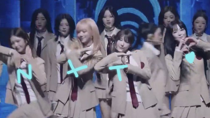 A group of female performers in school uniforms pose together on stage with their hands forming a heart shape, overlaid with the glowing cyan text "NXT" across the image, matching the post's caption.