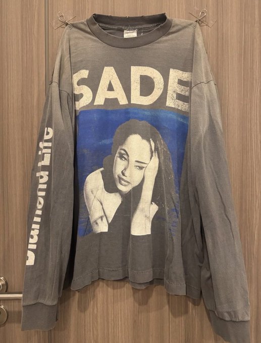 A gray long-sleeve graphic t-shirt featuring the word 'SADE' above a blue-toned portrait of the singer Sade, with 'smooth operator' printed vertically on the sleeve, hanging against a wooden door.