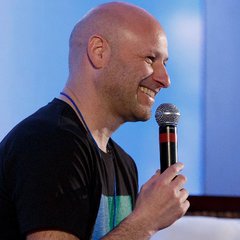 A bald man smiles while holding a microphone, captured mid-performance or speech. The image appears to be a stock photo of a speaker, unrelated to the accompanying post about Ethereum co-founder Joseph Lubin's massive ETH transfer.