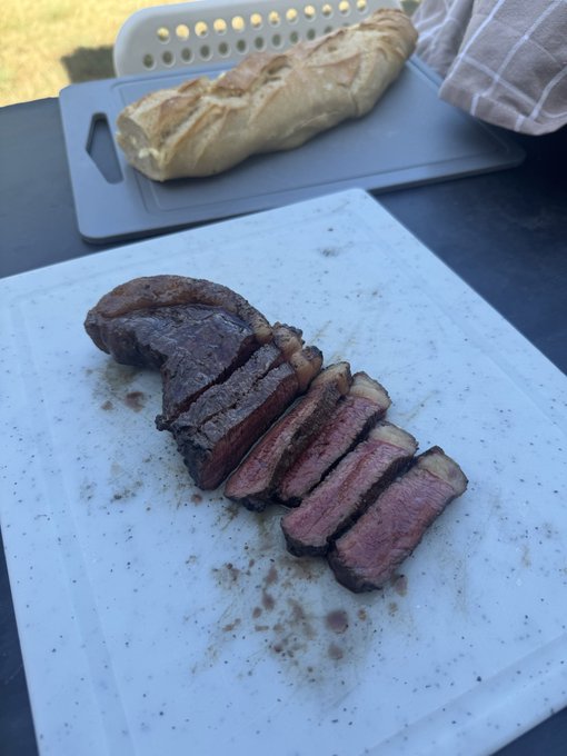 A sliced steak cooked to medium-rare, displayed on a white speckled cutting board, with a loaf of crusty bread on a separate cutting board in the background.