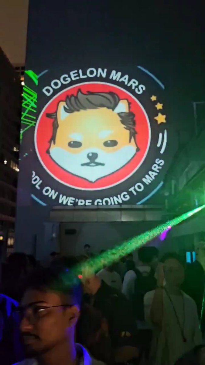 A large illuminated billboard displays "DOGELON MARS" with the tagline "LOL ON WE'RE GOING TO MARS," featuring a Shiba Inu dog illustration, while a crowd watches from below, tying directly to the $Elon (Dogelon Mars) memecoin promotion.