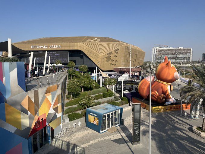 An outdoor daytime view of the Etihad Arena, featuring a large geometric building, colorful murals, and a giant inflatable orange cat sitting on a rooftop.