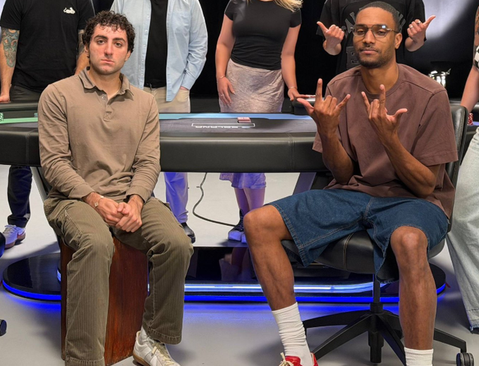 Two men sit in front of what appears to be a casino-style table with 'CYGNATRACK' visible; the man on the right makes hand gestures while wearing denim shorts, and the man on the left wears a tan polo.