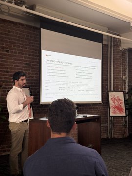 A man stands at a wooden podium giving a presentation, with a slide projected on a screen behind him titled 'Assessing the Future of...', in a room with exposed brick walls.