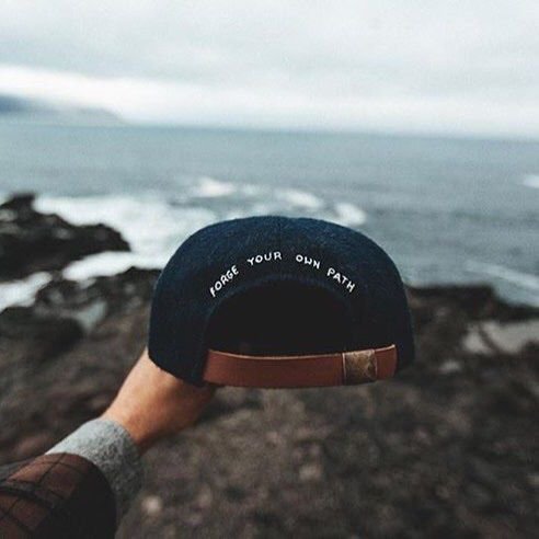 A hand holding up a dark navy baseball cap with the text 'FORGE YOUR OWN PATH' embroidered on the back, photographed against a coastal landscape with the ocean and rocky shore.