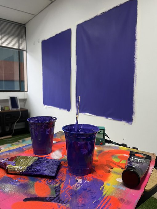 An artist's workspace showing two purple paint buckets, brushes, and a tube of paint on a colorful splattered table, with two purple-painted rectangular canvases hanging on the white wall behind.