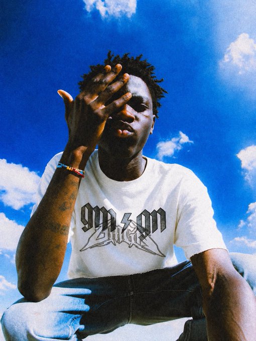 A photo of a person sitting against a blue sky with clouds, wearing a white t-shirt with 'YN OM' printed on it, partially covering their face with one hand.