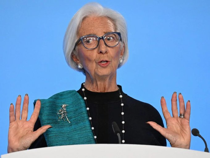 An older woman with white hair and glasses speaks at a podium, gesturing with both hands raised, wearing a teal jacket over a black top and a pearl necklace.