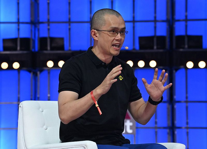 Changpeng "CZ" Zhao, founder of Binance, speaks animatedly at the Bitcoin Asia conference in Hong Kong, wearing a black polo shirt with the Binance logo while gesturing with both hands. The image visually supports the post's emphasis on his continued industry influence, capturing him mid-discussion on a blue-lit stage about crypto's infrastructure-focused future.