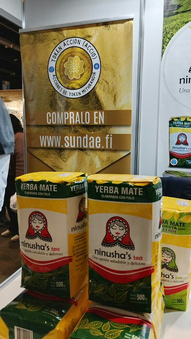 A promotional booth for Token ACCIÓN displays stacked packages of yerba mate, with a large sign reading “COMPRALO EN www.sundae.fi.” The tea boxes show “YERBA MATE,” “ninusha’s,” and “500 g,” while the post links the display to ACCIÓN and Cardano-related hashtags.