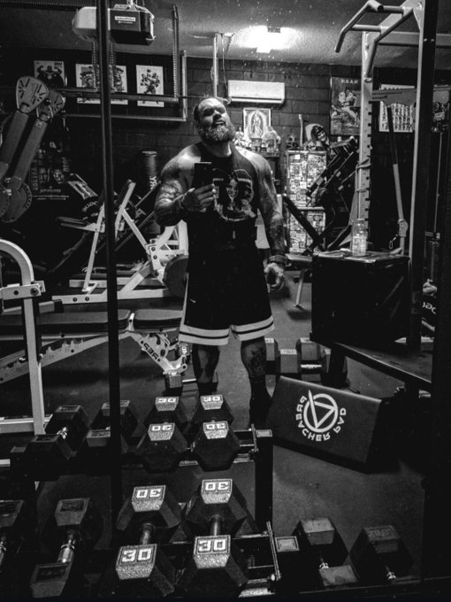 A bearded, tattooed bodybuilder stands in a home gym taking a mirror selfie with his phone, surrounded by rows of dumbbells labeled "30" and "20," with a "Synergy Crew" banner visible in the background. The image fits the post "Everyone about to get rekt" as the man's intimidating, muscular physique suggests he's ready to crush anyone who steps into his training space.