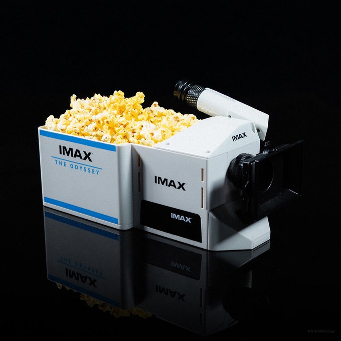 The image features a box of popcorn sitting next to a small video camera, both placed on a black surface. There are several pieces of popcorn visible inside the box, indicating that they are ready to be enjoyed by moviegoers or snack enthusiasts. The presence of the video camera suggests that this could be an advertisement for a movie or event featuring the popcorn.