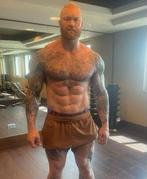 A muscular, heavily tattooed red-haired man wearing only shorts stands in a gym in front of a mirror.