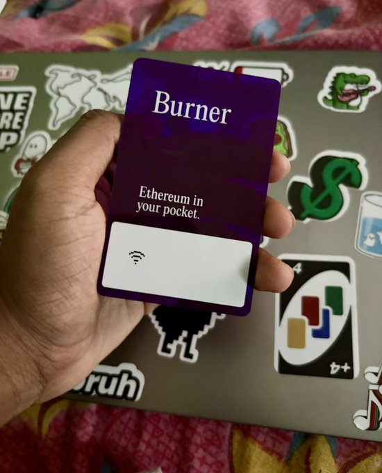 A hand holds up a purple "Burner" NFC wallet card with the tagline "Ethereum in your pocket," displayed against a laptop covered in stickers including a crocodile, dollar sign, and various tech-related designs. The image showcases the affordable, open-source hardware wallet mentioned in the post, highlighting its compact card-style design with a visible NFC contactless symbol.