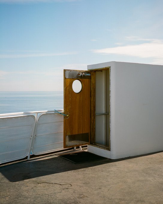 An open, weathered wooden door set within a white rectangular structure stands on a concrete pier overlooking the calm sea, symbolizing transition or the threshold between two worlds. The image visually represents the concept of "The Other Side of Open," now sold to @thisistolo for 0.2 ETH.