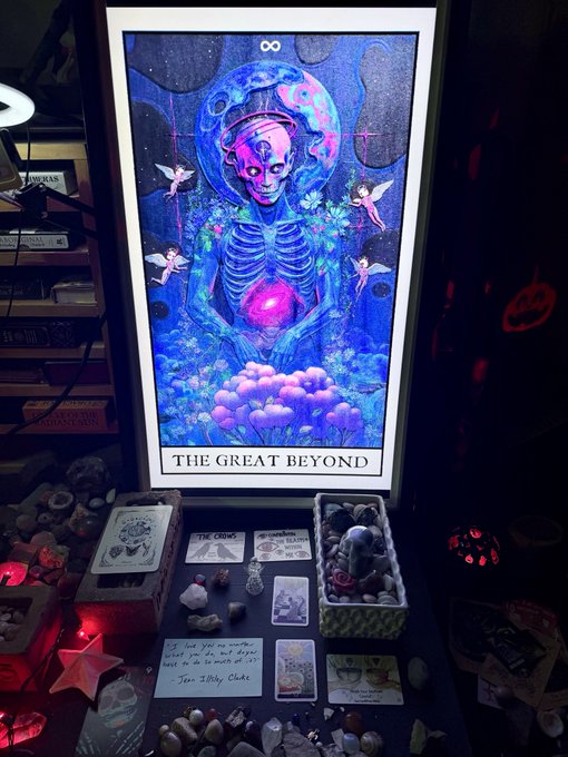 An illuminated poster reading 'THE GREAT BEYOND' depicts a skeletal alien figure surrounded by fairies and a glowing heart, displayed above a table arranged with tarot cards, crystals, and a quote card from Jen Satsune.