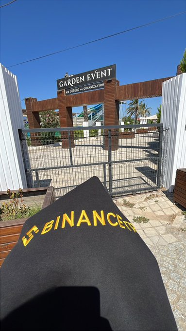 A view of a closed garden entrance with a sign reading "GARDEN EVENT - THE DINING by ORGANIZATION," with a piece of fabric bearing the text "LA BINANCEIT" in the foreground.