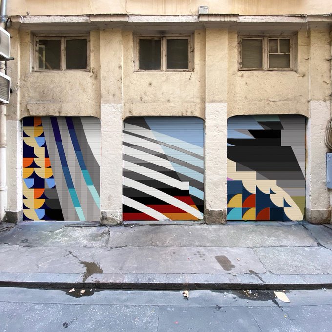 Three garage doors on a weathered building facade are painted with colorful geometric and abstract mural designs in shades of blue, yellow, white, black, and red.