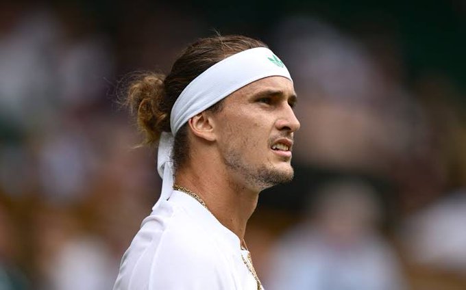 A side profile close-up of a tennis player with long hair tied back, wearing a white headband with a small green logo and a white shirt, with a gold chain visible around his neck, appearing to be at Wimbledon.