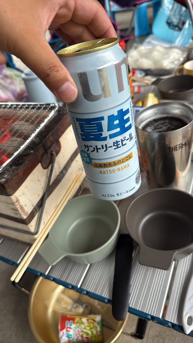 A hand holding a Japanese Suntory 'Utsunomiya Natsu-Nama' beer can at what appears to be a camping setup with cookware visible in the background.