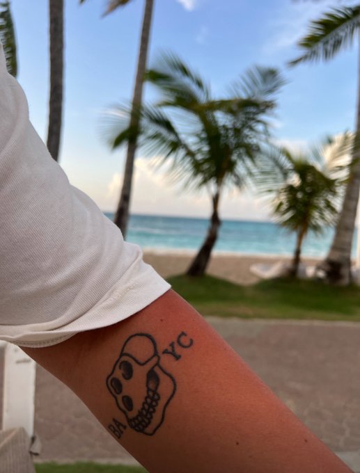 The image features a person's arm with a tattoo of a skull, along with the text "@BoredApeYC" written below it. The tattoo appears to be located on the inner forearm or elbow area. The background shows palm trees and a beach, suggesting that this location is near a coastal region. The post seems to be promoting a sense of community and connection among its followers by using the @BoredApeYC account as an example.