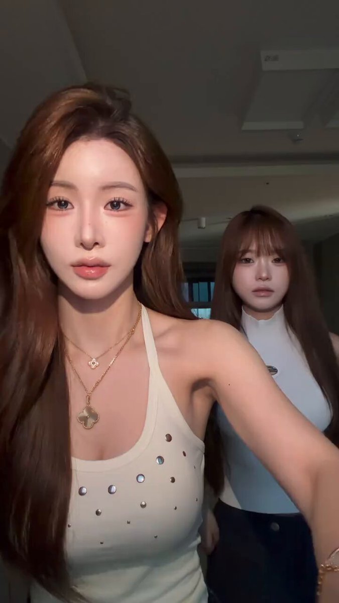 The image features two women standing next to each other, both wearing white tops with gold accents. The woman on the left has long brown hair and a necklace, while her companion on the right also has long brown hair but without any jewelry. They are positioned in front of a window, which adds depth to the scene. The background is blurred, drawing attention to the two women as the main focus of the image.