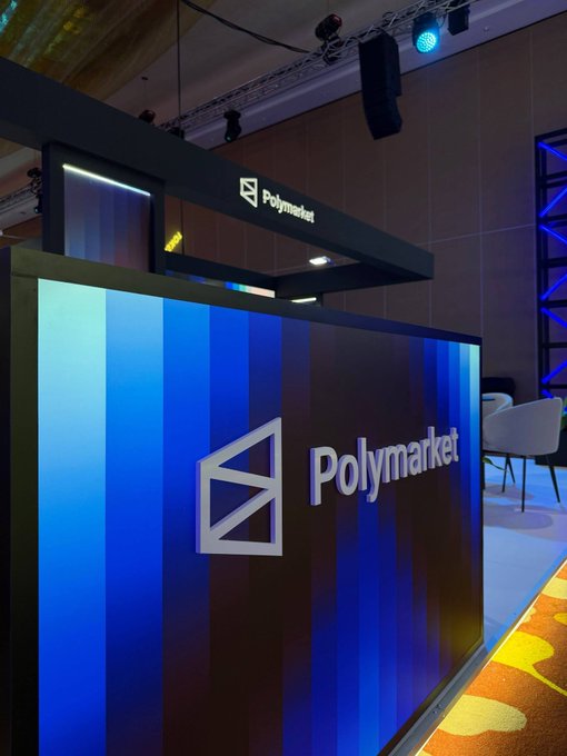 An indoor event space features a booth with a blue striped wall and the 'Polymarket' logo and name prominently displayed.