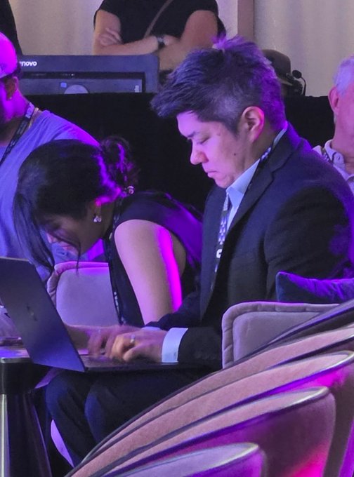 A man in a dark suit and a woman beside him are both intently looking down at a laptop screen in a dimly lit room with purple lighting.