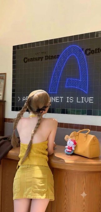 A woman with braided hair wearing a yellow dress and hair bows stands at a counter looking at a large LED sign reading 'Century Diner' and 'APP INTERNET IS LIVE'.