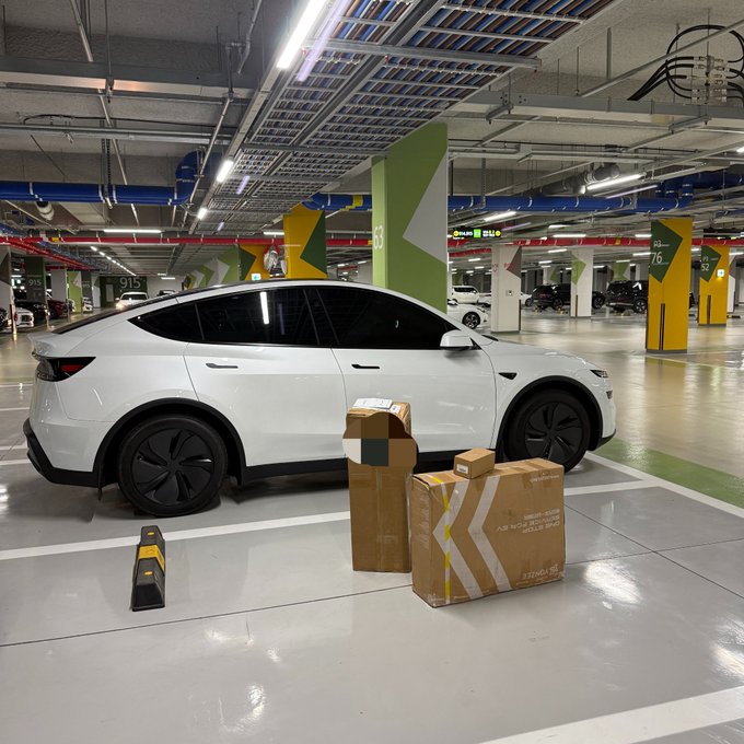 A white Tesla is parked in a numbered underground parking garage (spaces K-3 and G-6 visible on green and yellow pillars), with cardboard boxes placed beside it and a yellow-and-black wheel chock on the ground in front. The scene suggests the owner is preparing to swap in a new battery or component, aligning with the post's mention of a "2nd battery" addition to their Tesla.