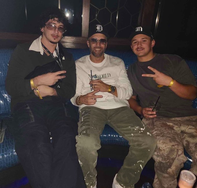 Three men sit together on a blue booth in a dimly lit venue; the man in the center wears a white 'No Worlds' sweatshirt and sunglasses, while the men on either side both wear black caps with a logo and make peace signs.