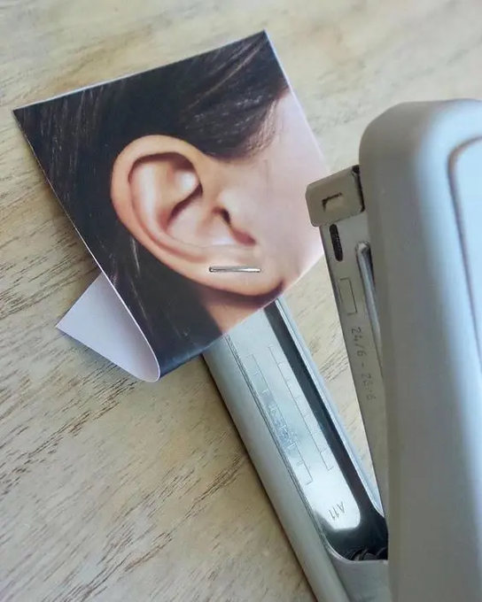A photograph showing a silver stapler resting on a piece of paper printed with an image of a person's ear, with a metal staple visible on the printed ear.