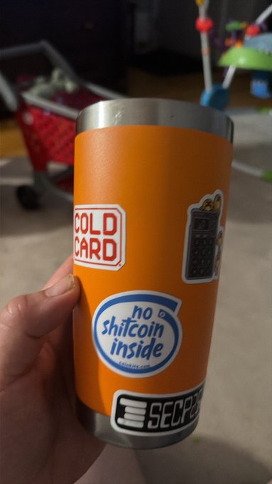 A hand holding an orange tumbler cup decorated with various stickers, including ones reading 'COLD CARD', 'no shitcoin inside', and 'SECP256'.