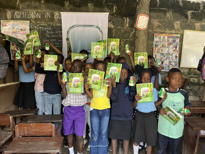 A group of children standing in a classroom holding up green packages and small bottles, with educational posters visible on the concrete walls behind them.
