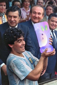 Diego Maradona smiling as he holds up an LG smartphone displaying a smiley face emoticon, with onlookers visible in the background.