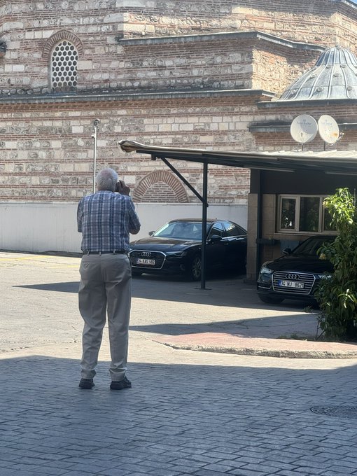 An older man in a blue plaid shirt and khaki pants stands on a brick-paved street, photographing a black Audi sedan parked near a brick building with a small dome.