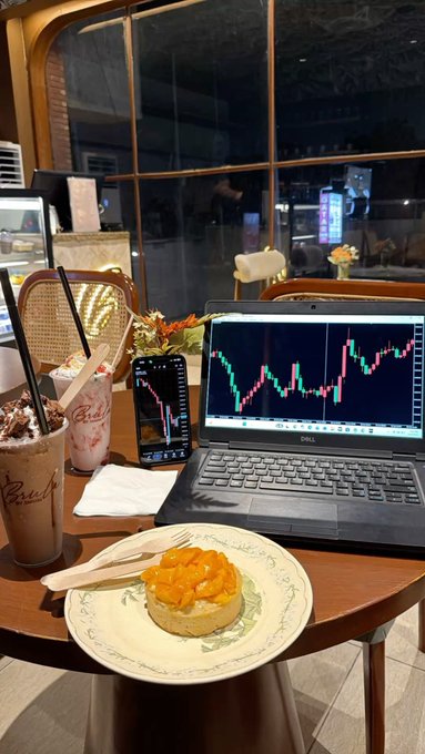A cafe table scene with a laptop and smartphone showing stock market charts, a coffee cup, an iced drink, and a small cake topped with orange slices on a plate.