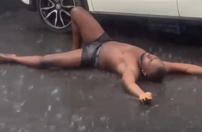 A man lies sprawled on a wet pavement next to a car, wearing only underwear, appearing to have fallen or collapsed on the ground.