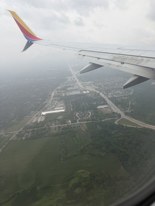 An aerial view from an airplane window showing the wing with red and yellow livery, overlooking a landscape of highways, buildings, and green fields below.