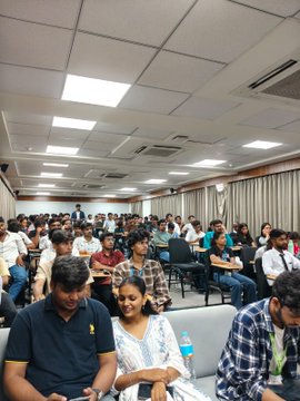 The image shows a large group of people sitting down and looking at their phones or tablets. The room appears to be a conference hall with rows of chairs and couches arranged for attendees. Some individuals are engaged in conversations, while others are focused on their devices. A few bottles can also be seen scattered around the room. The post features an image of a man wearing a yellow shirt and a woman sitting next to him, both looking at their phones or tablets.
