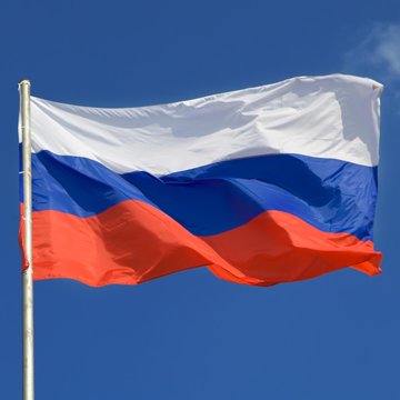The Russian flag with its white, blue, and red horizontal stripes waving in the wind against a clear blue sky.