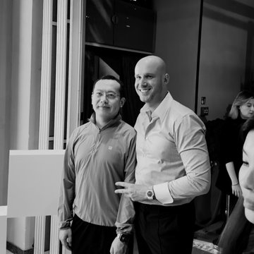 A black and white photo of two men posing together at an indoor event, one wearing glasses and a jacket, the other in a dress shirt with a watch.