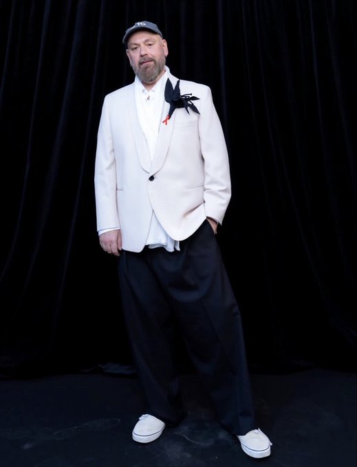 A bearded man wearing a "BOSS" cap stands confidently against a black curtain backdrop, dressed in a white blazer with a black lapel flower, a white shirt, and loose black trousers paired with white sneakers. The image reflects the post's message by showcasing someone proudly sporting their branded BOSS cap.