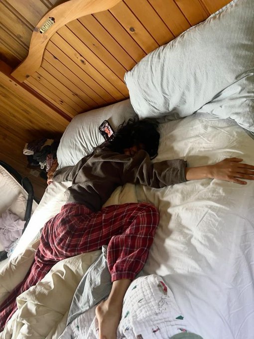A person with dark hair lies face down on a bed, wearing a gray top and red plaid pajama pants, with a phone resting nearby.