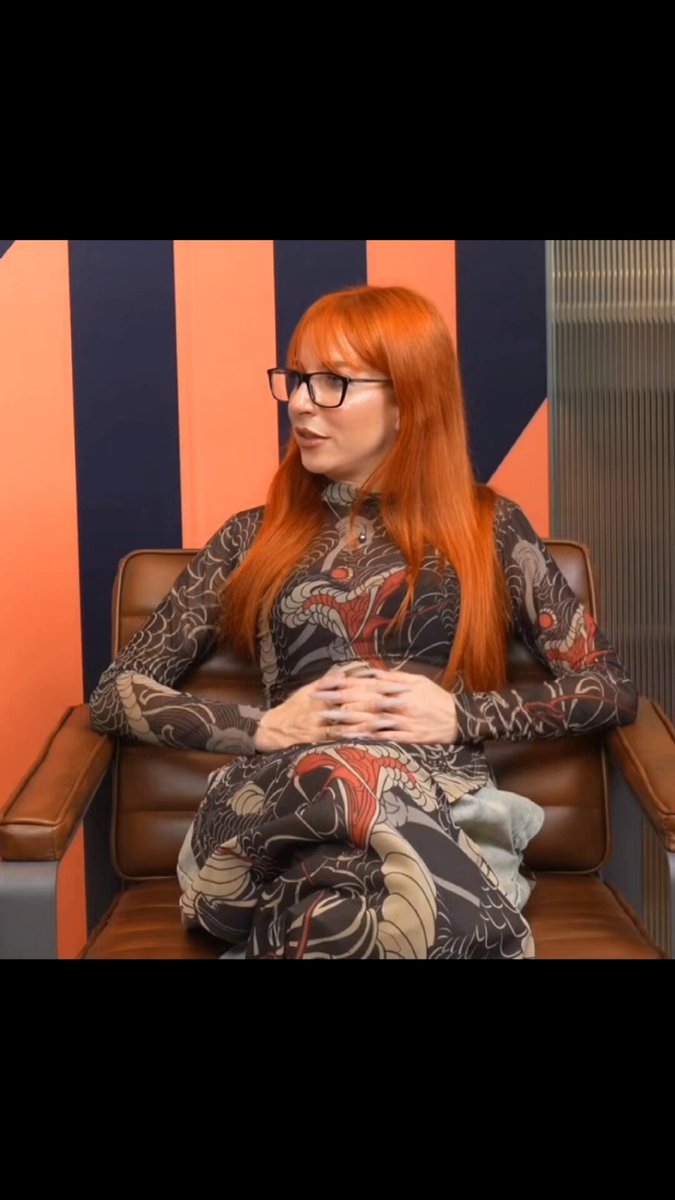 A woman with red hair wearing a dress with oriental patterns sits on an office chair in front of a desk. She wears glasses and looks away from the camera as she speaks or stares at something out of focus, possibly explaining her lack of interest in NFTs. The image also includes text that refers to the value of NFTs coming from floor price glennon, suggesting there is more to understanding it than just surface-level information.