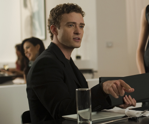 A young man in a black suit sits at a restaurant table, looking off to the side.