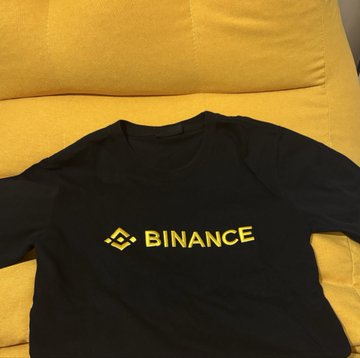 A black Binance cotton t-shirt with the yellow Binance logo and text displayed on the front lies on a yellow chair. The image showcases one of the items from the Binance swag giveaway bundle mentioned in the post.