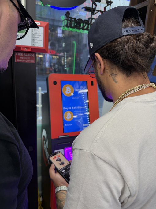 Two people stand at a Bitcoin ATM; one person in a white shirt and black cap holds a phone displaying an image while looking at the screen which shows 'Buy & Sell Bitcoin'.