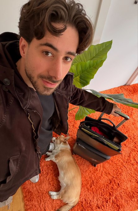 A young man in a brown jacket takes a selfie on an orange rug with a kitten playing near a small suitcase and a potted plant.